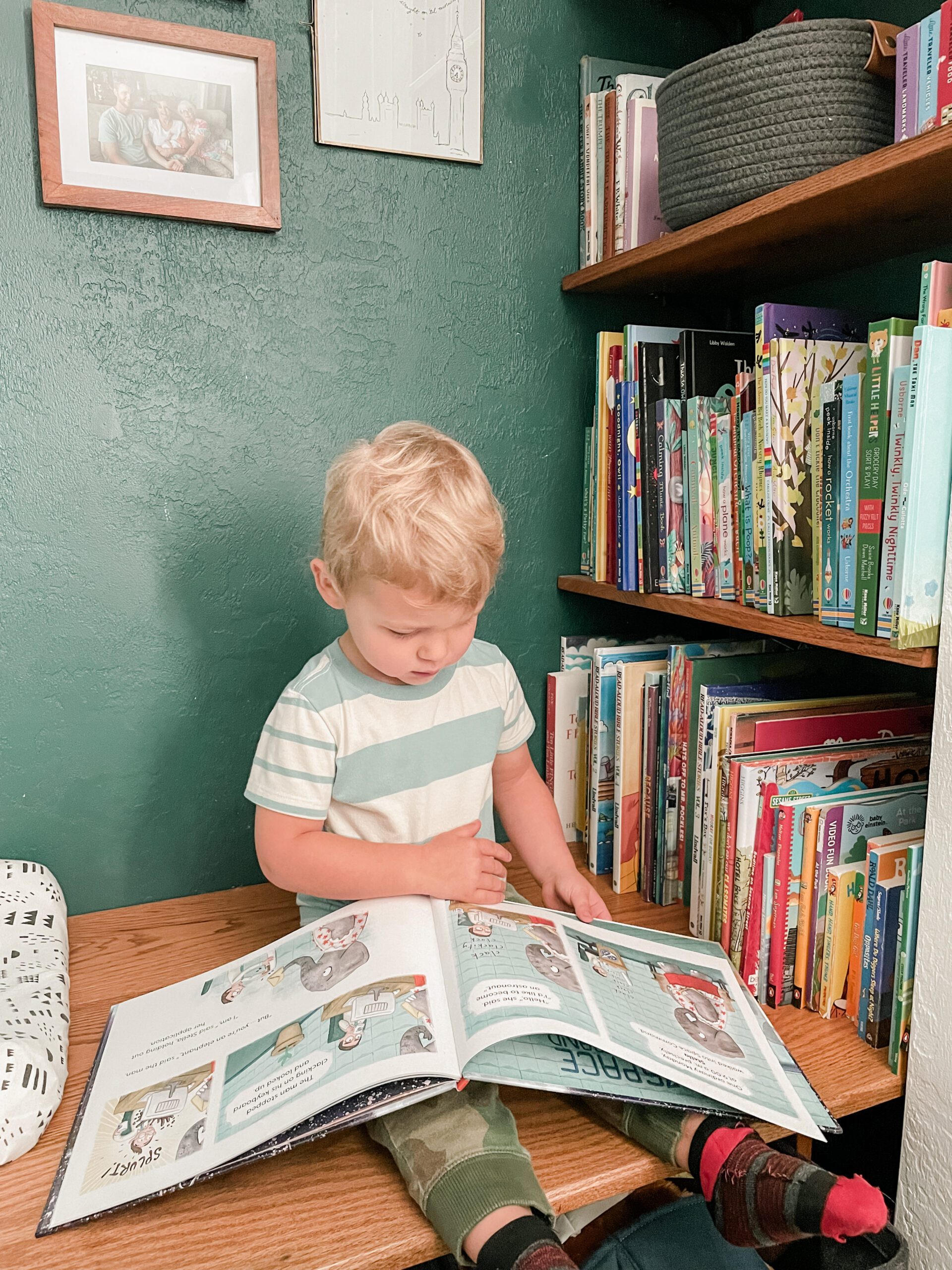 Little boy reading inside of closet area converted into a reading nook. He is sitting on a bench with a book on his lap. Shelves filled with books are behind him.