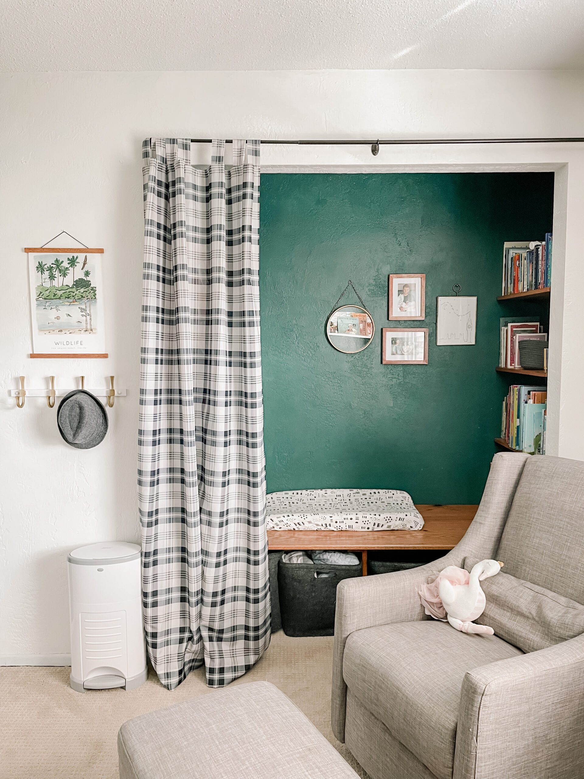 Image of closet that is painted green. It includes shelves filled with books, a bench with a changing pad, baskets below the bench, pictures and a mirror hanging on the wall. Area is sectioned off with a black and white plaid curtain.