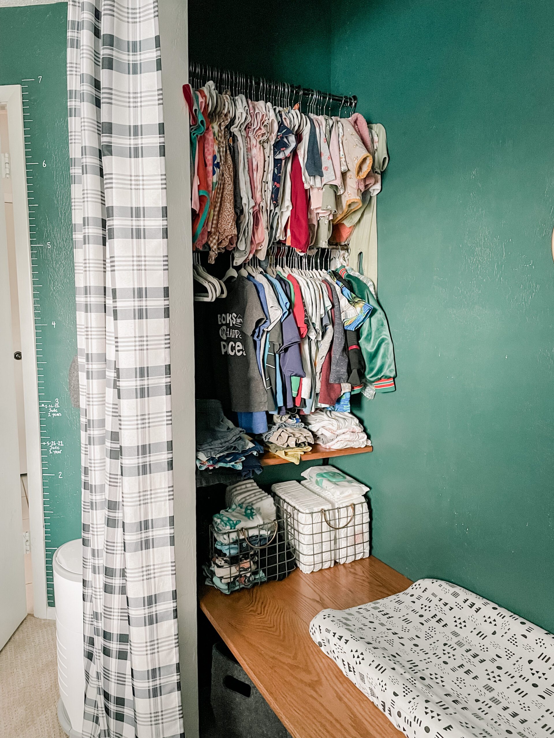 Image of inside corner of a closet. Two tension rods hold children's clothing. Baskets filled with diapers. Area sectioned off with a black and white plaid curtain