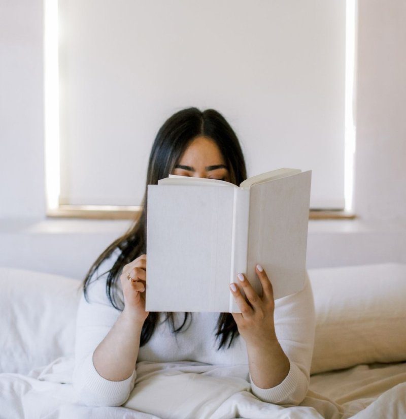 A woman sits on a bed, engrossed in reading a book, surrounded by a cozy and serene atmosphere