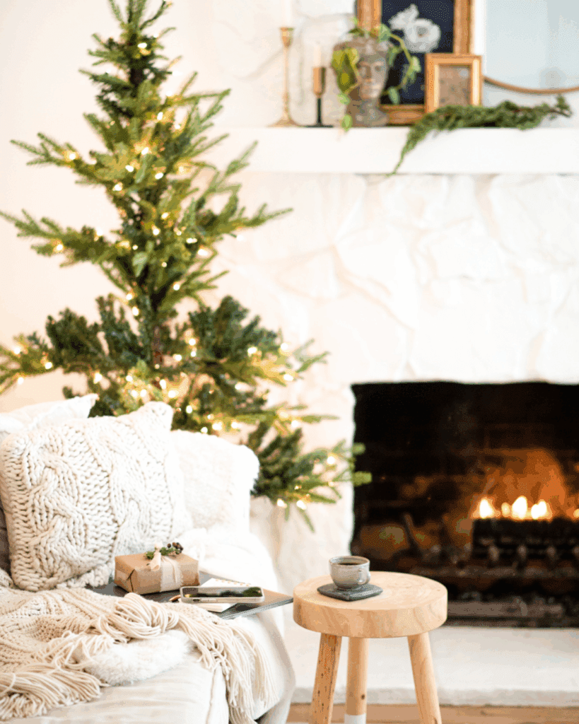 A Christmas tree beside a fireplace. Cozy blankets and pillows on a couch with a coffee and a little gift from the Holiday gift guide for women who love to read.