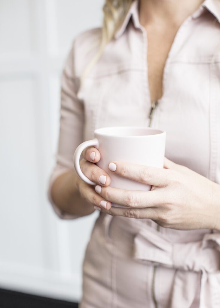 Image of a woman (see her neck to her waist) wearing light pink jumpsuit. She is holding a light pink coffee cup.