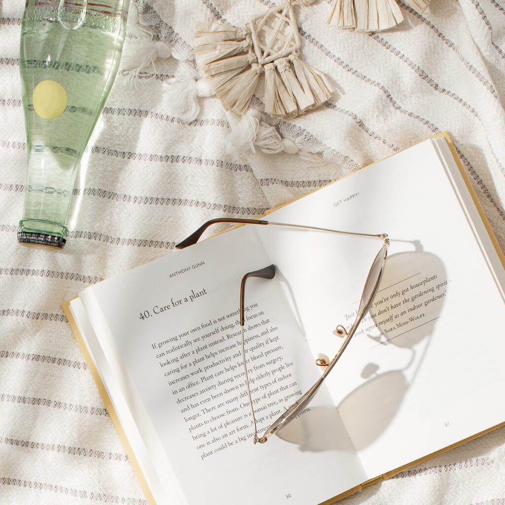 Summery image with a tan, striped blanket in the background. Open book on the blanket with sunglasses resting on top. A glass water bottle in the upper-right corner.
