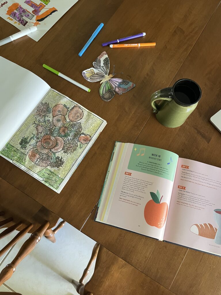 Flatlay image of the table with the book open on it. You also see crafts, coloring books, and markers, and a cup of coffee.